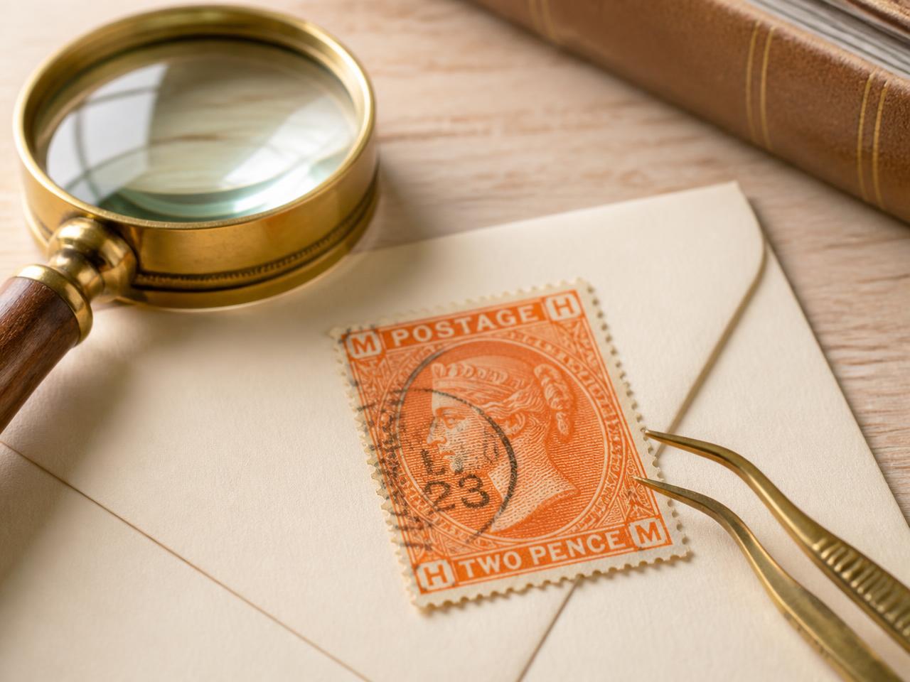 A vintage orange postage stamp on a cream envelope beside a magnifying glass and stamp tweezers